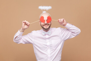 Portrait of funny bald bearded man with nimb over head, standing covering his eyes with little red hearts, hiding, wearing light pink shirt and bow tie. Indoor studio shot isolated on brown background