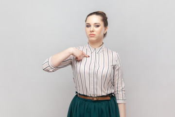 Portrait of strict bossy serious woman wearing striped shirt and green skirt standing pointing at herself with proud expression, looks arrogant. Indoor studio shot isolated on gray background.