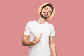Portrait of satisfied proud bearded man in white T-shirt and hat standing pointing at himself, looking at camera with optimistic confident expression. Indoor studio shot isolated on pink background.