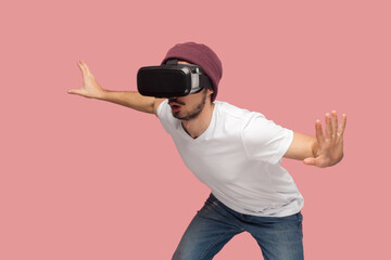 Portrait of amazed wary bearded man in white T-shirt and beany hat, wearing vr, playing in headset,...