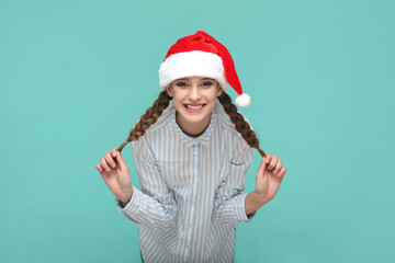 Portrait of funny optimistic teenager girl with braids wearing striped shirt and Santa Claus hat, looking at camera and pulling her pigtails aside. Indoor studio shot isolated on green background.