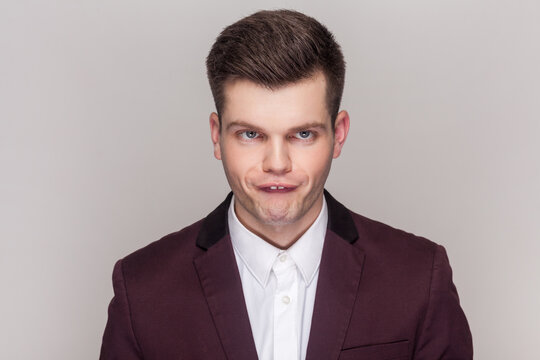 Portrait of crazy comic foolish handsome young man standing looking at camera with funny crossing eyes, wearing violet suit and white shirt. Indoor studio shot isolated on grey background.