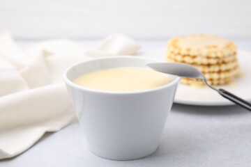 Tasty condensed milk in bowl and spoon on light grey table, closeup