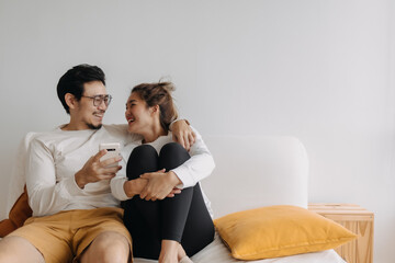 Happy lovely asian couple sharing smartphone to watch together in living room. - obrazy, fototapety, plakaty