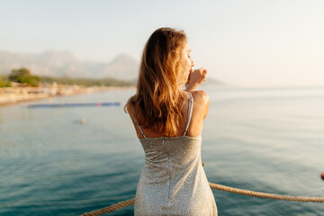 A slender girl with long hair in a gray shiny dress walks along the pier by the sea at sunset. A beautiful woman in an evening dress poses against the backdrop of the ocean at dawn. Summer background