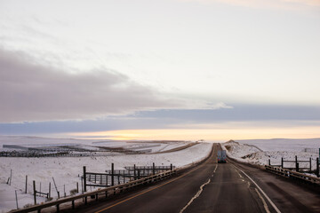 Fototapeta premium Beautiful winter landscape with a highway between blue snowy mountains, fluffy clouds on a sunset orange sky. Winter snow background. Elk Mountain, Wyoming, USA. Picturesque winter sky