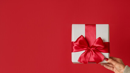 Holiday surprise. Romantic greeting. Female hand showing wrapped gift in white box with ribbon bow isolated on red copy space background.