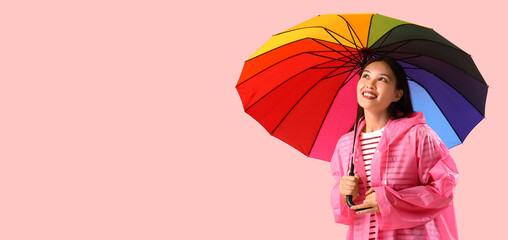 Beautiful young Asian woman in raincoat holding rainbow umbrella on pink background with space for...