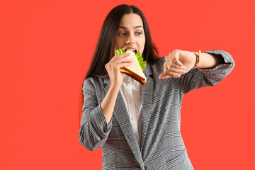 Young businesswoman eating tasty sandwich and looking at wristwatch on red background