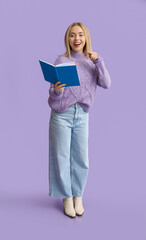 Beautiful young happy woman with book pointing at viewer on lilac background