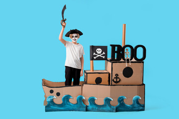 Little boy dressed for Halloween as pirate with cardboard ship on blue background