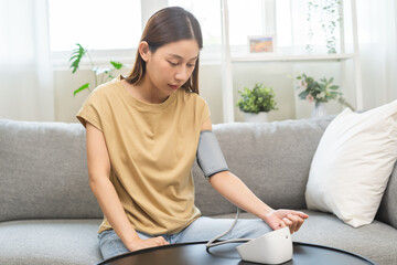 Hypotension problem asian young woman sitting on couch checking high blood pressure and heart rate with digital pressure monitor, making self check up pulse on arm, cuff at home. Healthcare medical. - obrazy, fototapety, plakaty