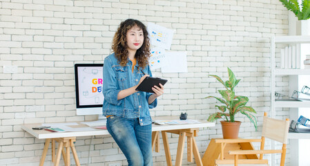 Asian professional successful young female creative graphic designer in casual fashionable denim jeans jacket standing smiling holding tablet computer working in workspace in company office.