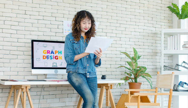 Asian professional successful young female creative graphic designer in casual fashionable denim jeans jacket standing smiling holding colors chart working in workspace in company office.