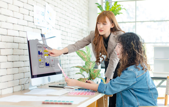 Asian professional successful young female creative graphic designer and colleague in casual fashion suit smiling helping choosing sampling comparing artwork on computer moniter in office workplace