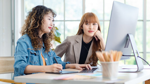 Asian professional successful young female creative graphic designer in casual outfit sitting smiling drawing sketching artwork with stylus pen when colleague talking chit chat taking break in office