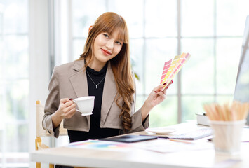 Asian professional successful young female businesswoman creative graphic designer in casual fashionable suit outfit sitting smiling choosing colors sample from pattern chart when taking coffee break