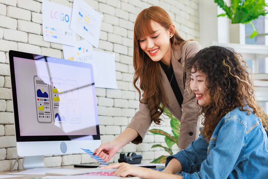 Asian professional successful young female creative graphic designer and colleague in casual fashion suit smiling helping choosing sampling comparing artwork on computer moniter in office workplace