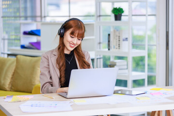 Fototapeta premium Asian professional successful young female businesswoman creative graphic designer in casual outfit sitting smiling wearing headphone listening to music playlist while working typing laptop computer