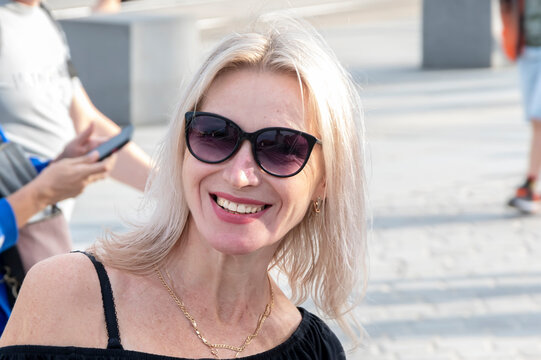Portrait of a happy blonde 45-50 year old woman wearing sunglasses on a neutral background of a European city.