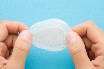 Young adult woman hand fingers holding and showing white new sticking plaster for eye covering on light blue table background. Pastel color. Top down view. Closeup.