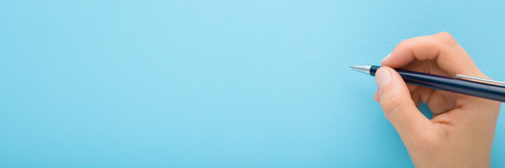 Young adult woman hand holding pen and writing on light blue background. Pastel color. Closeup. Empty place for text. Wide banner. Top down view.