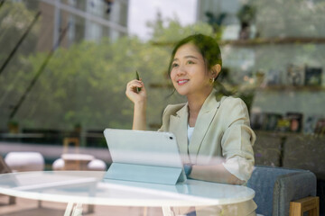 Beautiful smart Asian businesswoman is using a tablet and thinking while looking outside at a cafe.