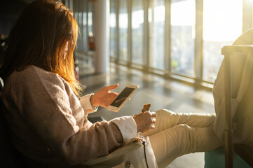 Unrecognizable woman using smartphone and eating while sitting with luggage in airport waiting room