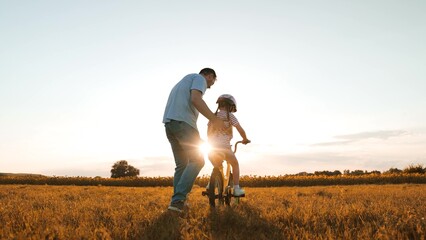 Caring father helps little daughter to ride bicycle in evening meadow on weekend