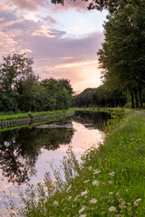 Fototapeta na wymiar A captivating scene of a dramatic purple-pink sky with beautifully articulated clouds, bathed in the setting sun's glow, mirrored in the tranquil waters of a serene river or canal. Dramatic Purple