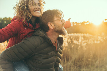 Happy couple have fun together in outdoor leisure activity in nature field during sunset time and golden hours light. Man carry woman on his back. People enjoying life and laughing. Winter autumn day - obrazy, fototapety, plakaty