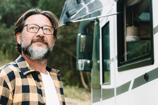 One mature adult man standing outside a modern rv recreational vehicle motorhome and looking around nature and parking destination. Concept of alternative lifestyle vanlife and vacation travel. People