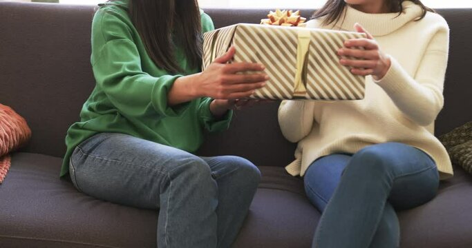 Midsection of biracial mother and adult daughter exchanging christmas gift on couch, slow motion