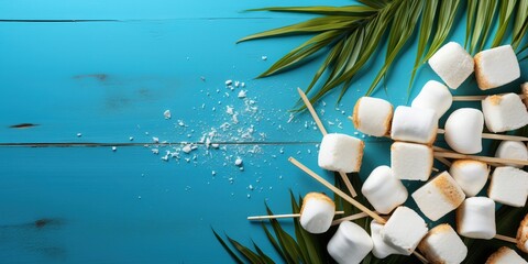 Food composition with white marshmallow on wooden sticks and palm leaves on a blue background with copy space. Summer vacation banner with traditional testy snacks for American open air picnic - obrazy, fototapety, plakaty