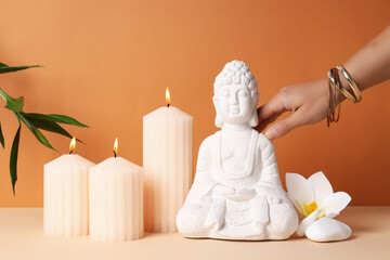Buddha statue, candles, flower and hand with bracelets on orange background
