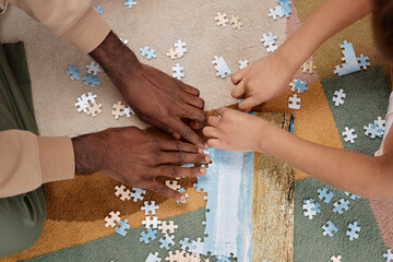 Top view closeup of father and kid solving jigsaw puzzle together on floor at home, focus on hands holding pieces