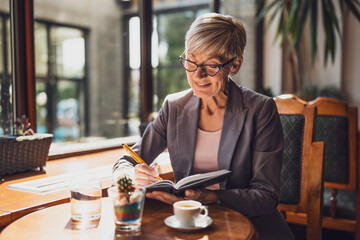 Mature woman is sitting in cafe and relaxing. She is drinking coffee and using note pad.