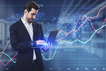 Attractive young businessman using laptop with creative business graph with index and grid on blurry office interior background. Stock market and financial statistics concept. Double exposure.