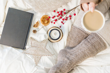Cozy winter scene. Christmas vibes. Woolen socks. Woman having coffee in bed with book and candle.