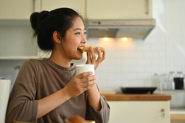 Happy young asian woman eating a slice of bread with jam for breakfast and drinking coffee in kitchen