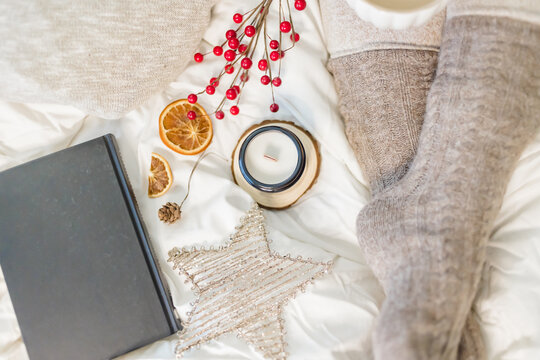 Cozy winter scene. Christmas vibes. Woolen socks. Woman having coffee in bed with book and candle.