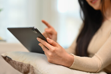 Cropped shot of young woman in warm sweater using digital tablet on couch at home