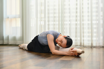 Fototapeta na wymiar Young ballerina dancer stretching leg on wooden floor in ballet studio