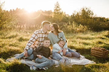 Mother, father, daughter, son sitting on blanket, hugging in grass in field at sunset. Family...
