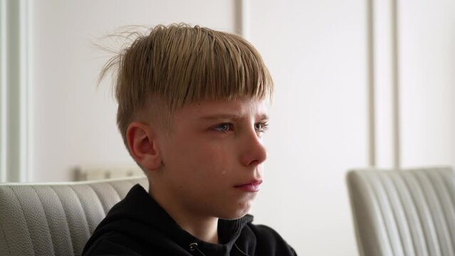 Frustrated teen boy sits on chair in the living room and lets a tear out of his eye