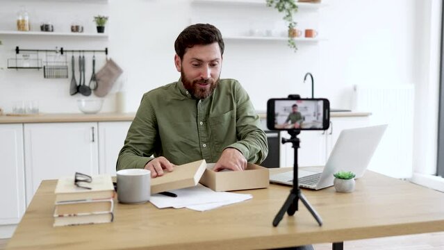 Positive handsome man recording video blog while unboxing new modern smartphone indoors. Interesting influencer leaving review about product and sitting at table with laptop in room.