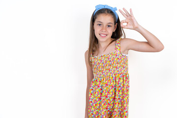 Beautiful kid girl wearing floral dress hold hand arm okey symbol toothy approve advising novelty news