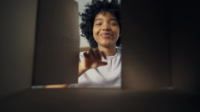 Young happy african american woman open cardboard box and looks at toothy smile with delight. Home lifestyle. Bottom view at portrait cute lady. Concept unpacking, product delivery and movement.