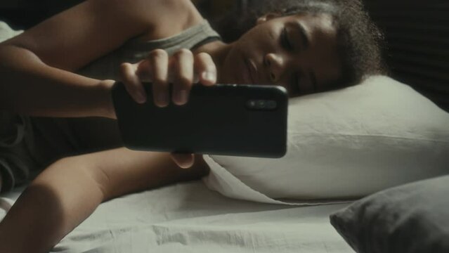 Sad teen girl lying on bed at night, receiving message on smartphone, reading it and getting upset about cyberbullying