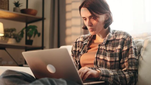 Portrait of pretty young woman freelancer having distance remote work typing browsing scrolling products in internet online store on laptop computer and eating cookie from pocket at light home indoors